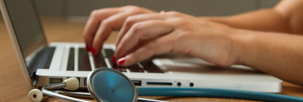 Woman typing on laptop at desk with coffee mug, representing online health resources and virtual functional medicine consultations